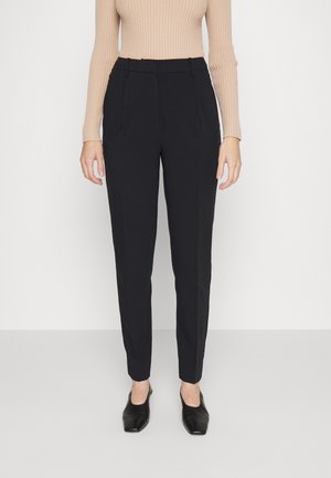 Pantaloni neri su misura con una vestibilità dritta, caratterizzati da pieghe frontali e passanti per cintura, abbinati a un top beige a coste e scarpe nere.