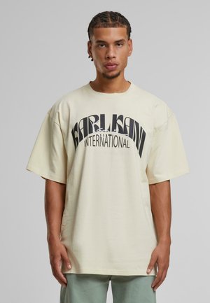 Cream oversized T-shirt with "KARL KANI INTERNATIONAL" in bold black text. Short sleeves, round neck, and soft cotton texture.