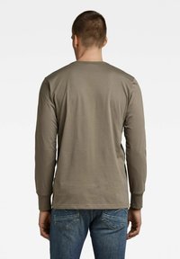 Mężczyzna w brązowej koszuli z długim rękawem i niebieskich dżinsach, widziany od tyłu na tle gładkiego białego tła.