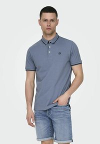 Camiseta polo gris con un cuello y ribetes negros, con tres botones y un pequeño logotipo. Combinada con pantalones cortos de mezclilla azul claro con dobladillo enrollado.