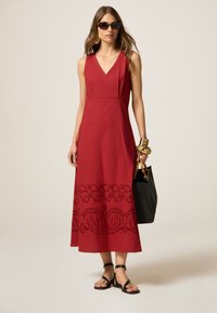 Rotes ärmelloses Maxikleid mit V-Ausschnitt, das am Saum mit filigranen schwarzen Stickereien verziert ist; getragen mit schwarzen flachen Sandalen und einer Tasche.