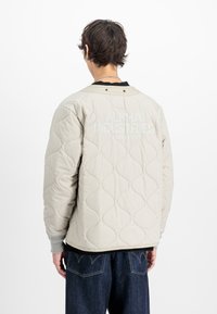 Persona, kas valkā gaiši beidzas, quilto apvalku ar tekstu "Alpha Industries Studio Edition" uz muguras, apvienojumā ar tumši ziliem džinsiem.