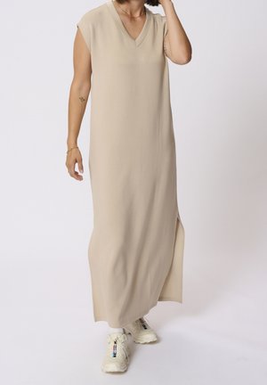 Vrouw draagt een mouwloze beige maxi-jurk met een zijsplit en witte chunky sneakers tegen een effen witte achtergrond.