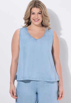 Femme souriante aux cheveux blonds ondulés vêtue d'un haut sans manches bleu clair et d'un pantalon assorti, debout devant un fond blanc uni.