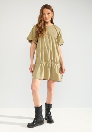 Donna con capelli lunghi che indossa un vestito beige largo a maniche corte e stivali neri fino a metà polpaccio, in piedi davanti a uno sfondo chiaro e semplice.