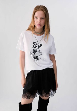 Junge Frau mit langen Haaren, die ein weißes Minnie-Maus-T-Shirt, schwarze Shorts mit Spitzenbesatz und schwarze kniehohe Stiefel vor einem einfarbigen Hintergrund trägt.