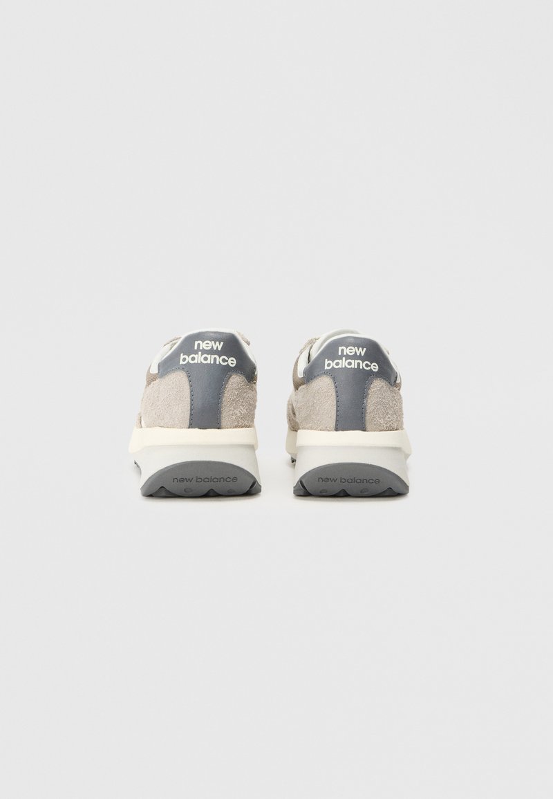 Graue und beigen Wildledersneaker mit einer weißen Zwischensohle, die ein auffälliges "New Balance"-Logo an der Ferse und ein elegantes Design aufweisen.