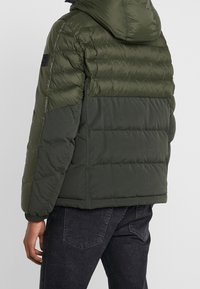 Mand iført en tofarvet olivengrøn polstret jakke med hætte og mørke jeans, vist fra ryggen mod en ensfarvet lys baggrund.