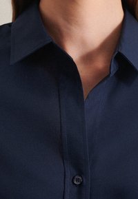 Närbild av en person som bär en knappad mörkblå skjorta med krage, som visar halslinjen och övre bröstområdet.