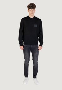 Maglione nero a maglia con collo rotondo e un patch logo testurizzato sul petto, abbinato a jeans scuri e sneakers bianche.