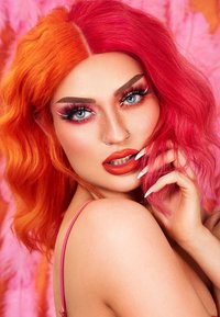 Capelli ondulati vibranti arancio e rosa, trucco audace con occhi blu sorprendenti, una carnagione liscia e unghie lunghe e bianche, il tutto su uno sfondo colorato.