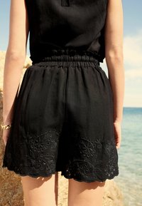 Les shorts en lin noirs disposent d'une taille élastique, d'un ourlet à volants et de motifs floraux brodés le long du bas.