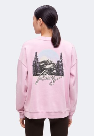 Femme aux cheveux foncés en queue de cheval portant un sweat-shirt rose avec une montagne, des arbres, le soleil et le script "Roxy" dans le dos.