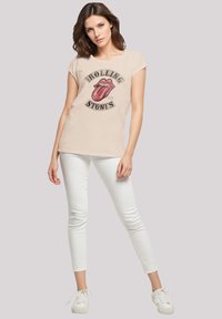Beige T-shirt with a red tongue logo and "ROLLING STONES" text. Paired with white skinny jeans and white sneakers. Short sleeves, relaxed fit.