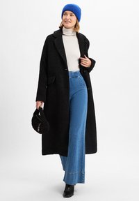 Schwarzer strukturierten Mantel, weiße Rollkragenjacke, blaue weit geschnittene Jeans und eine gestrickte blaue Mütze. Hält eine kleine schwarze Pelztasche.