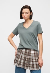 Νέα γυναίκα με κοντά σκούρα μαλλιά, φορά γκρι μπλουζάκι με V λαιμόκοψη πάνω από καρό καφέ και λευκή φούστα και μπλε τζιν, όρθια με το χέρι στη μέση.