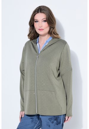 Frau mit einem olivgrünen Kapuzenpullover mit Reißverschluss und kleinen weißen Flecken, darunter ein blaues Hemd mit Kragen, stehend vor einem weißen Hintergrund.