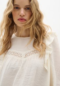 Blusa bianca leggera con colletto arricciato e bordo in pizzo ricamato. Texture morbida con un design ampio e fluido.