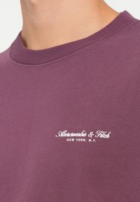 Lilla dressipluus pehmest puuvillast, millel on klassikaline ümmargune kaelus ja valge logo, mis kannab kirjut "Abercrombie & Fitch New York, N.Y."
