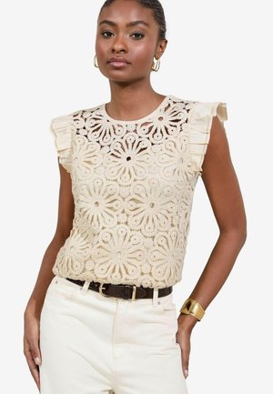 Top en dentelle couleur crème avec des découpes florales, manches courtes à volants et col rond, associé à un pantalon blanc et une ceinture noire.