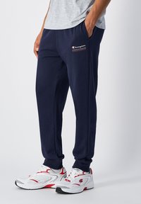 Pantalones de chándal azul marino hechos de una tela suave, con un logotipo en la pierna izquierda, bolsillos laterales y un corte relajado. Combinados con zapatillas blancas y rojas.