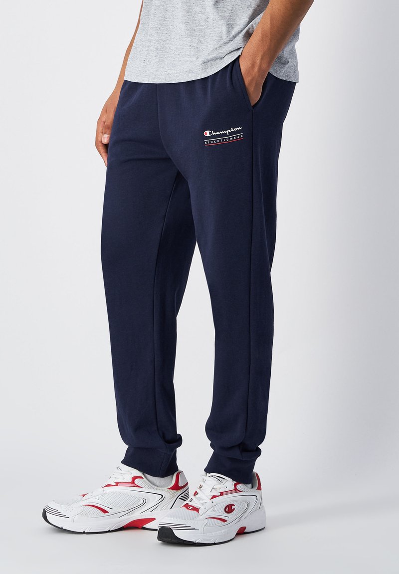 Pantalones de chándal azul marino hechos de una tela suave, con un logotipo en la pierna izquierda, bolsillos laterales y un corte relajado. Combinados con zapatillas blancas y rojas.
