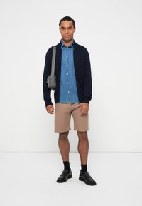 Marine cardigan over en denimskjorte, lysebrune ribbede shorts, sorte sokker og slip-on sko. Bærer en liten grå veske over skulderen.