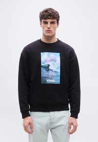 Νεαρός άνδρας που φοράει μαύρο φούτερ με γραφικό γαλάζιο κύμα ωκεανού και κείμενο "HUGO", συνδυασμένο με ανοιχτόχρωμο τζιν.