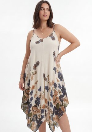 Femme portant une robe crème sans manches avec un imprimé floral bleu et marron et un ourlet asymétrique, debout avec une main sur la hanche.