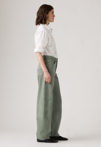 Femme aux cheveux bruns mi-longs se tient de côté, portant une chemise blanche à manches longues, un pantalon large vert sauge et des ballerines noires sur un fond uni.