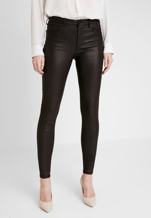 Woman wearing shiny black skinny pants, white long-sleeve blouse, and beige pointed-toe high heels, standing against a white background.