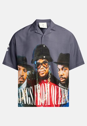 Camicia a maniche corte con bottoni, caratterizzata da una stampa grafica di tre figure, prevalentemente nei toni blu, grigio e rosso, con testo bianco.