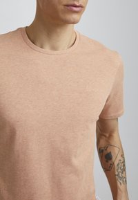 Tricou din bumbac de culoare piersică, cu guler rotund și mâneci scurte. Prezintă un logo mic pe piept și un material moale, texturat.