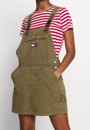 Osoba nosząca oliwkową sukienkę ogrodniczkową z przednią kieszenią oznaczoną Tommy Jeans oraz czerwoną i białą koszulkę w paski z krótkim rękawem.