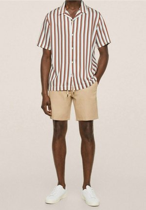 Mies, joka käyttää lyhythihaista nappipaitaa, jossa on pystysuorat ruskeat ja valkoiset raidat, beiget shortsit ja valkoiset tennarit.