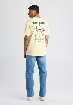 Geel katoenen t-shirt met een grafisch ontwerp op de achterkant, featuring een platenspeler, wijnglas en tekst. Gecombineerd met blauwe jeans en witte sneakers.