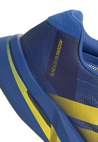 Blauer und gelber Sportschuhabsatz mit Mesh-Struktur, gepolstertem Kragen und der Aufschrift „Adizero Boston 13“ auf der Seitenfläche.