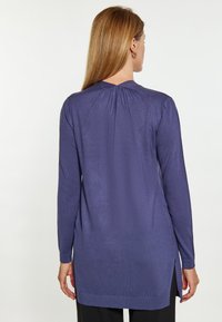 Camiseta de manga larga en púrpura intenso, con un escote trasero fruncido, puños acanalados y aberturas laterales para mayor movimiento. Textura suave.