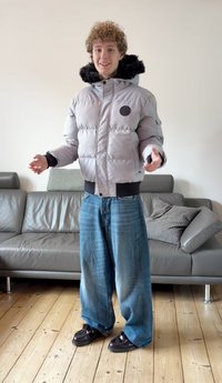 Szürke puffertalpú dzseki szőrmével bélelt kapucnival, cipzáras zsebbel és bordázott mandzsettákkal. Széles szabású kék farmernadrág és fekete cipő. Világos fa padló háttérrel.