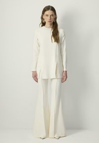 White, long-sleeve tunic paired with matching flared pants; features side slits, smooth texture, and a clean, minimal design.