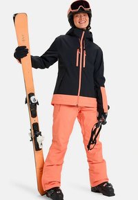 Person in schwarzer und pfirsichfarbener Skiausrüstung, die pfirsichfarbene Ski und Skistöcke hält, einen Helm und eine Skibrille trägt und lächelt.