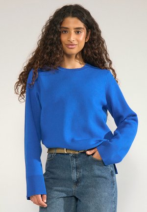 Jeune femme aux longs cheveux bouclés portant un pull bleu vif rentré dans un jean bleu taille haute avec une ceinture beige.