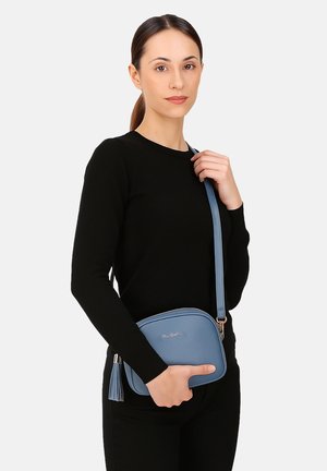 Borsa a tracolla azzurro chiaro con forma arrotondata, materiale testurizzato, con un accento di nappina e tracolla regolabile, tenuta da un modello in un maglione nero.