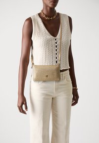 Persona che indossa un gilet senza maniche a trecce color crema, pantaloni color crema, collana in oro, bracciale in oro e una piccola borsa a tracolla in oro con texture.