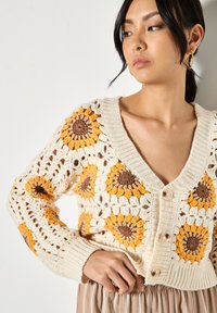 Cárdigan de crochet con patrón de girasol; base crema con detalles en amarillo y marrón; cuello en V profundo, cierre de botones, puños y dobladillo acanalados.