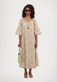 Frau trägt ein beiges, blumiges Midikleid mit Puffärmeln, gelbe Sandalen, Sonnenbrille, Creolen und hält eine hellgrüne Handtasche.