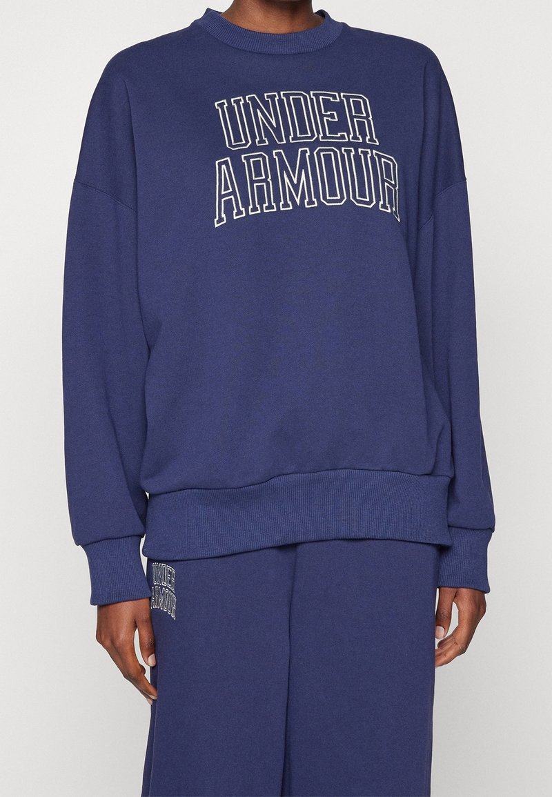 Sudadera oversized de color azul marino con puños y dobladillo acanalados, que presenta el logo "UNDER ARMOUR" en blanco a lo largo del pecho. Textura de tela suave.
