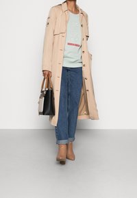 Persona que lleva un abrigo trinchera beige, un sudadera azul clara con texto, jeans azules con dobladillo, botas de tobillo color miel, sosteniendo un bolso negro y beige.