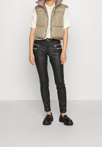 Chaleco acolchado en verde oliva, llevado sobre un polo beige claro, combinado con pantalones ajustados negros con detalles de cremallera plateada y mocasines negros.