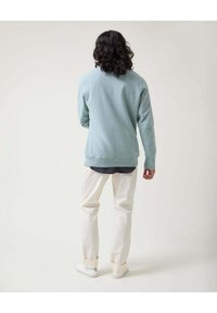 Sudadera azul claro con puños y dobladillo acanalados, llevada sobre una camisa oscura, combinada con pantalones color crema y zapatillas blancas.